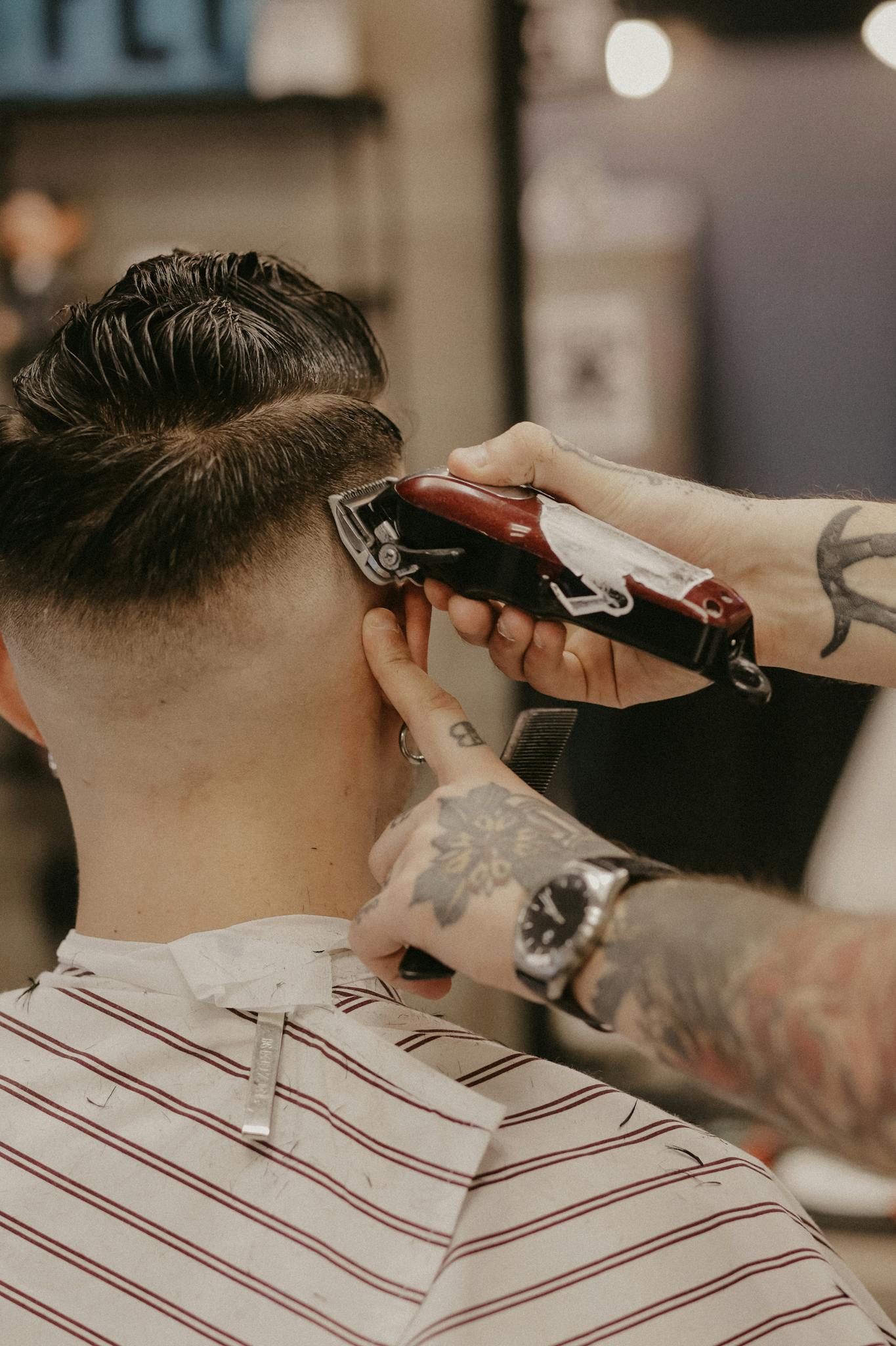 A tattooed barber uses electric clippers for a stylish haircut on a customer in a barber shop.